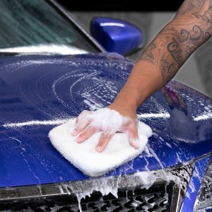 Técnico da SPA Window Film realizando esfregação técnica com luva de microfibra e shampoo Meguiar's em veículo azul.